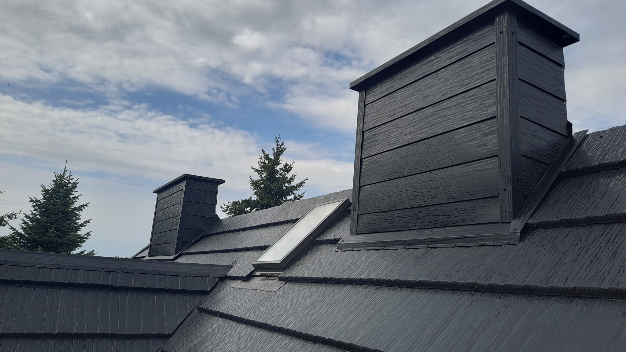 Czarny dach z blachy trapezowej z dwoma kominami wykończonymi deską elewacyjną i oknem dachowym, na tle błękitnego nieba z chmurami i zielonych drzew.