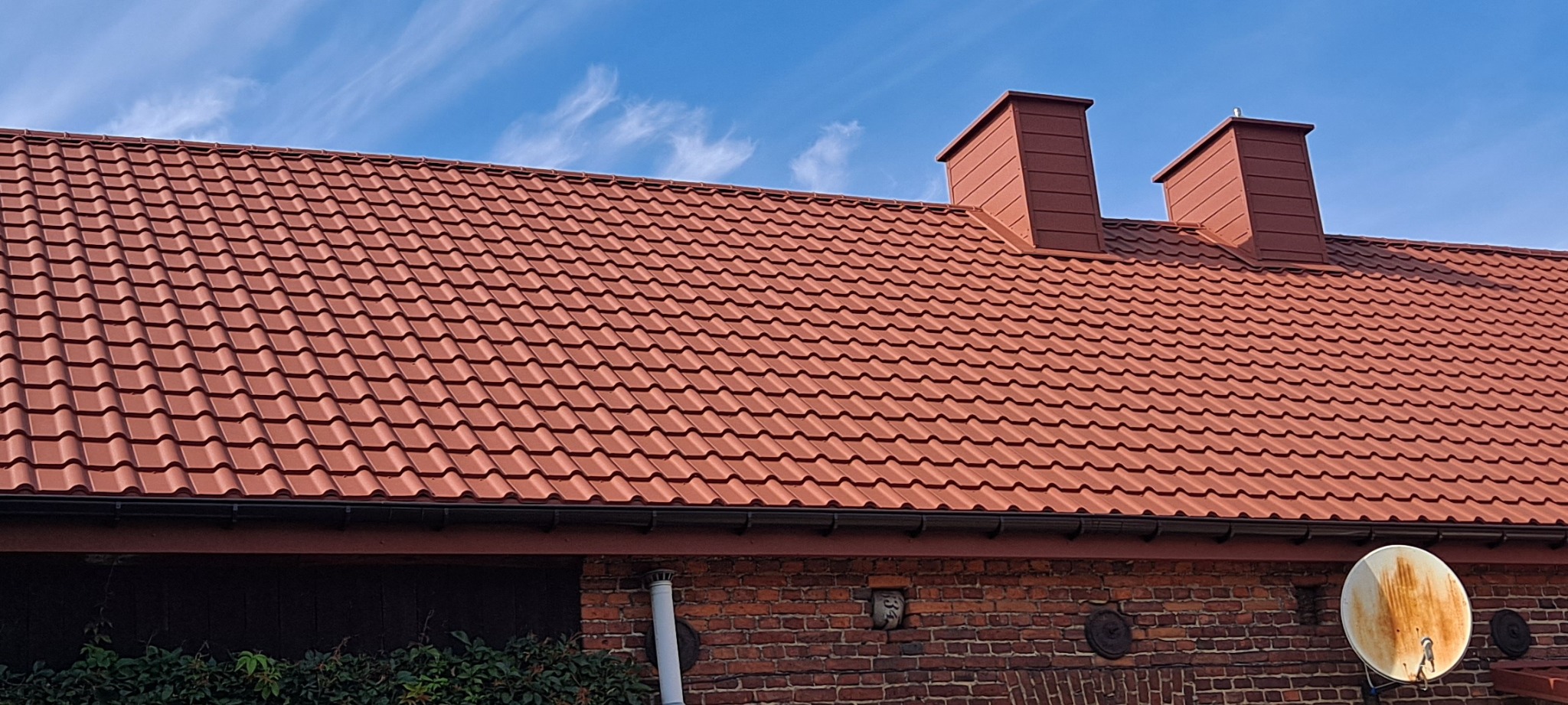 Czerwony dach z dachówki ceramicznej, dwa kominy w kolorze dachu, czarne rynny, ceglana ściana budynku z anteną satelitarną.