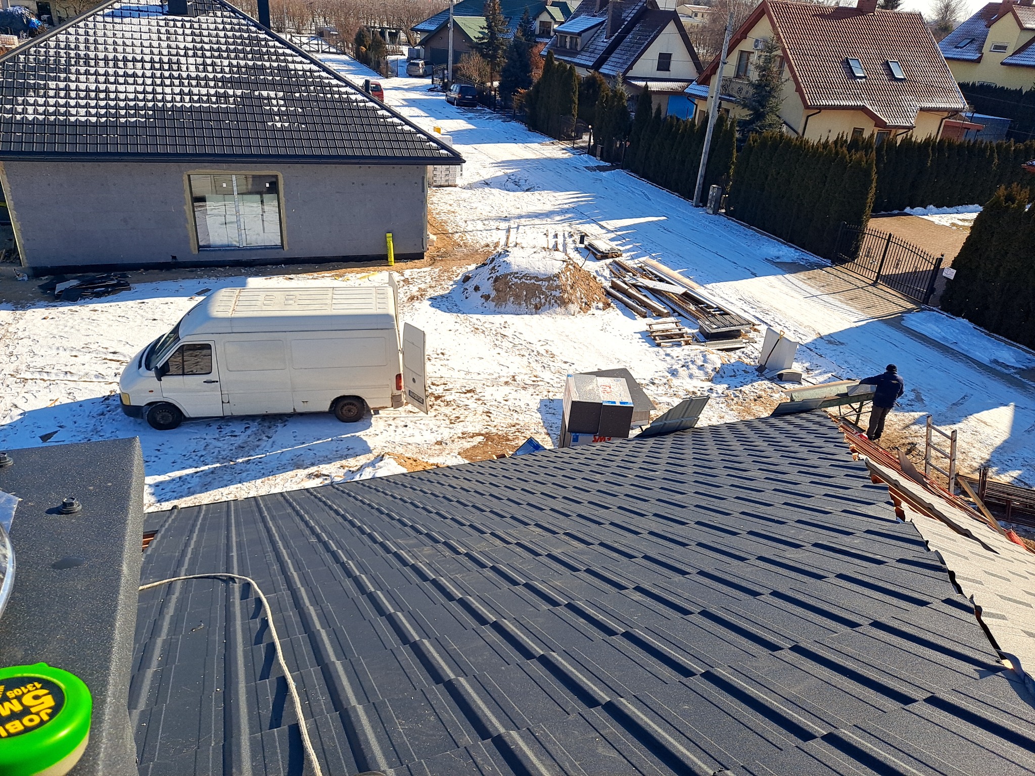 Nowy, grafitowy dach z blachy trapezowej w trakcie montażu, widok z góry na częściowo pokryty dach, w tle biały samochód dostawczy i zasypana śniegiem ulica z zabudowaniami.