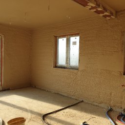 Daniel Piórek - Wnętrze pokoju po nałożeniu tynku strukturalnego na ściany i sufit, z widocznymi oknami zabezpieczonymi folią i elementami instalacji.