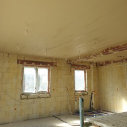 Daniel Piórek - Wnętrze w trakcie prac tynkarskich: ściany z pustaków, częściowo otynkowane, dwa okna z folią ochronną, widoczne instalacje i narzędzia, sufit z nałożonym tynkiem.