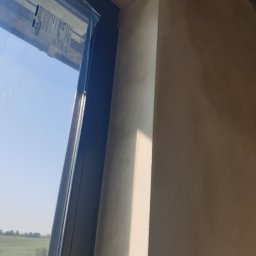 Daniel Piórek - Gładka, beżowa ściana po tynkowaniu, widoczna obok okna z widokiem na zielone pola. Świeżo położony tynk z delikatną fakturą w naturalnym świetle.