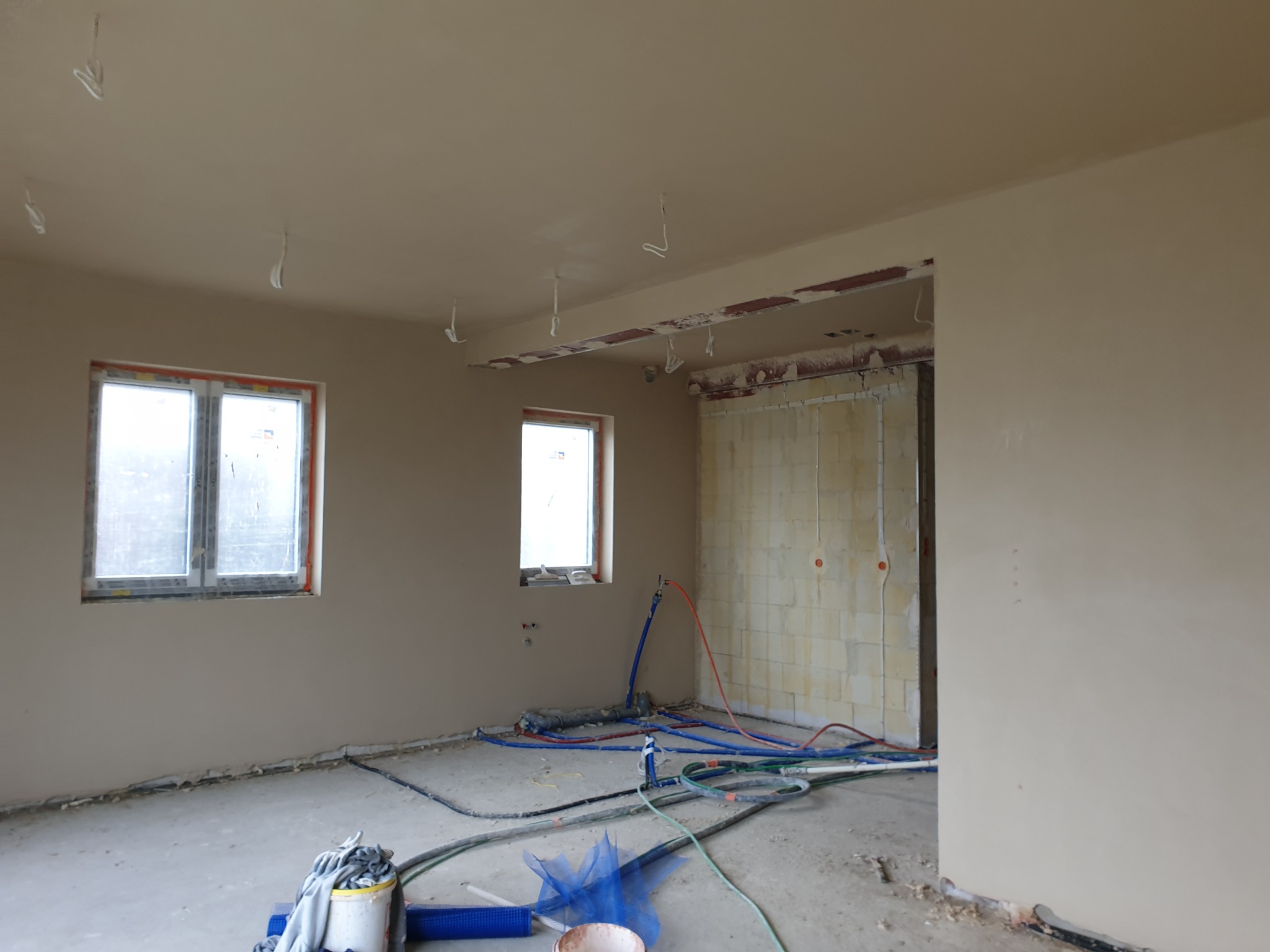 Wnętrze po tynkowaniu: gładkie ściany w odcieniach beżu, dwa okna, instalacje na podłodze, przygotowane do dalszych prac wykończeniowych. Widoczne pozostałości po pracach.
