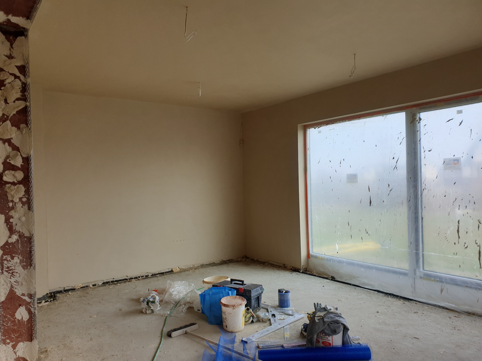 Pokój po nałożeniu jasnych tynków, z widocznymi narzędziami i materiałami budowlanymi, okno zabezpieczone folią. Częściowo otynkowany narożnik.