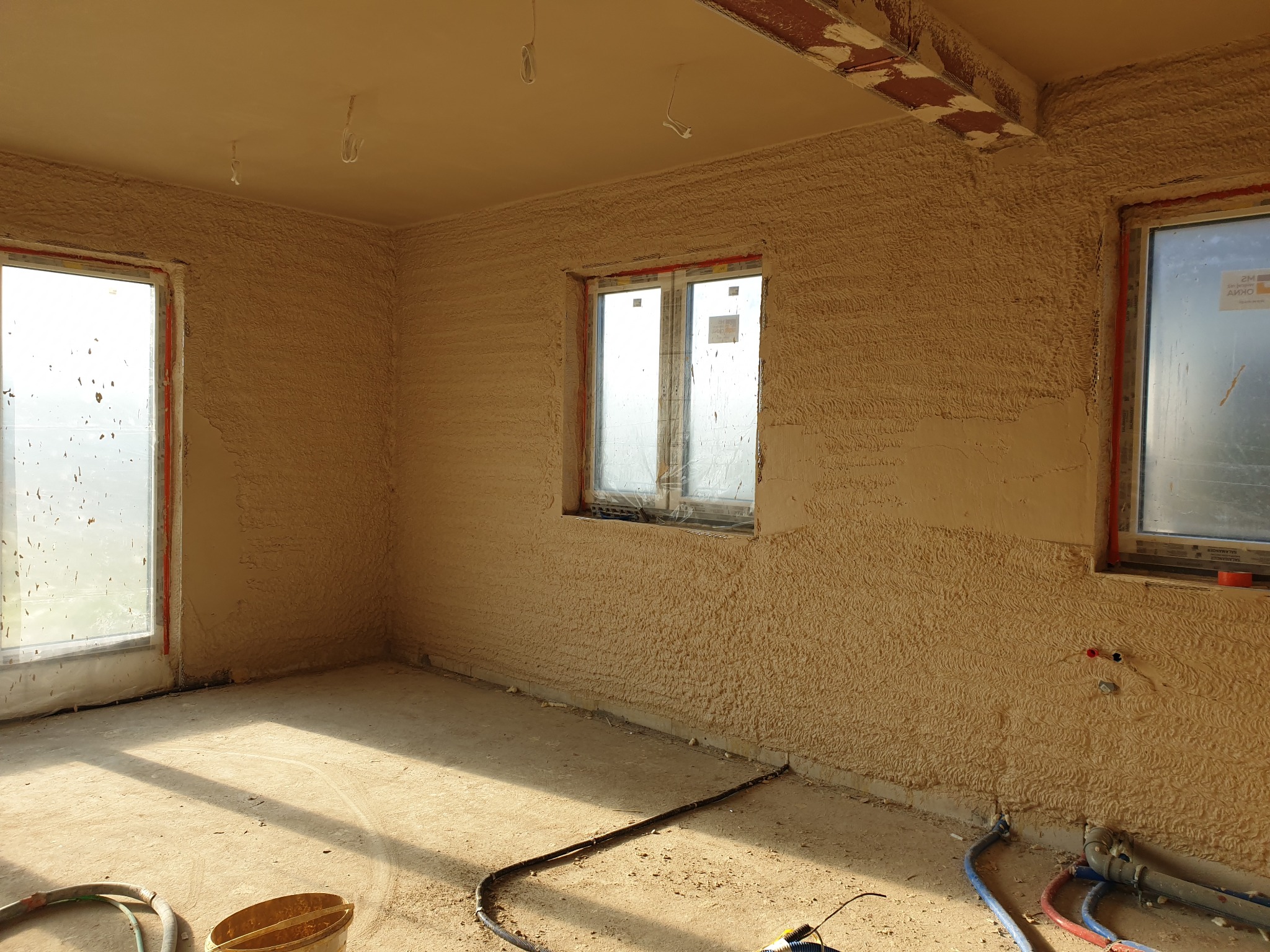 Wnętrze pokoju po nałożeniu tynku strukturalnego na ściany i sufit, z widocznymi oknami zabezpieczonymi folią i elementami instalacji.