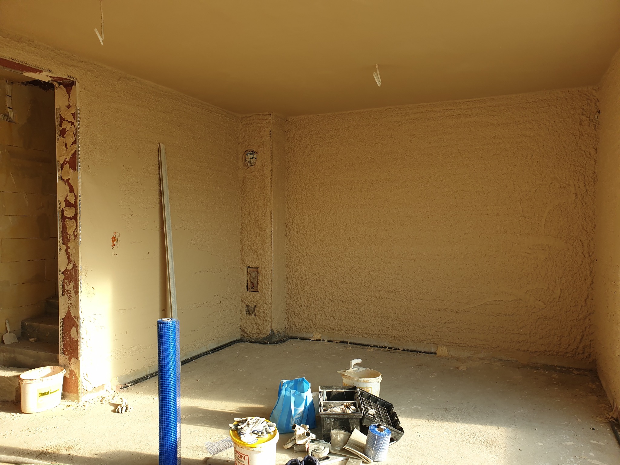 Pokój po tynkowaniu maszynowym: ściany i sufit pokryte świeżym tynkiem, widoczne narzędzia i materiały budowlane. W tle niezabezpieczone przejście do innego pomieszczenia.