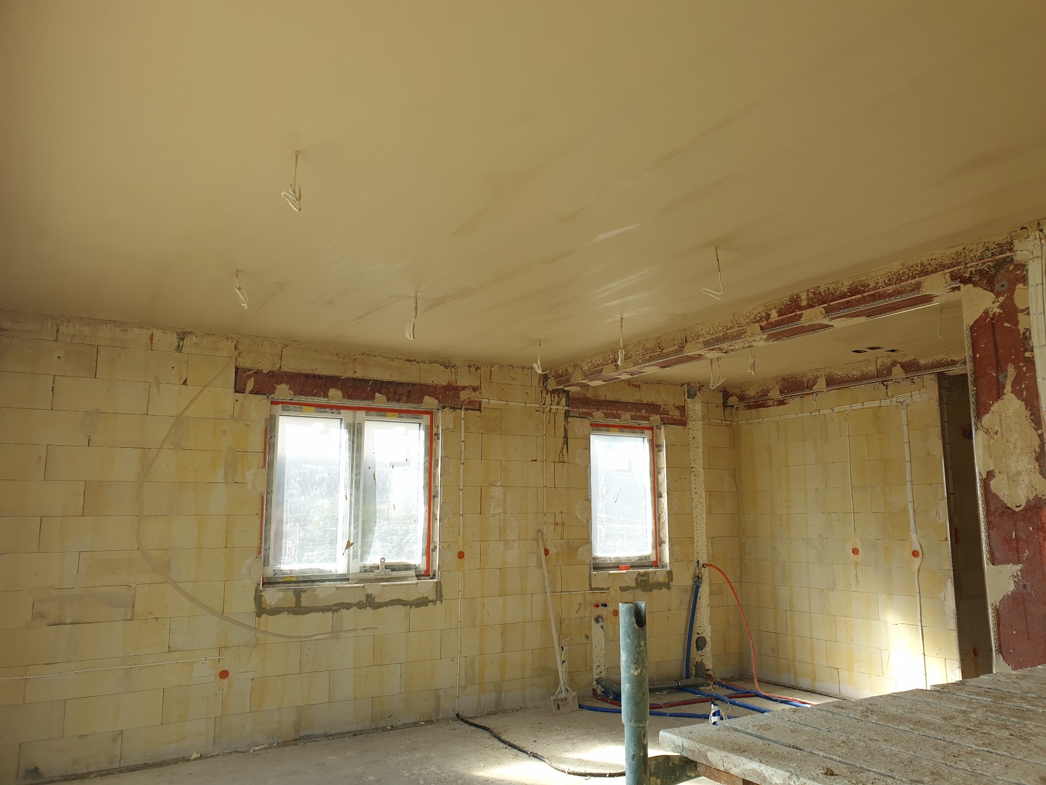 Wnętrze w trakcie prac tynkarskich: ściany z pustaków, częściowo otynkowane, dwa okna z folią ochronną, widoczne instalacje i narzędzia, sufit z nałożonym tynkiem.