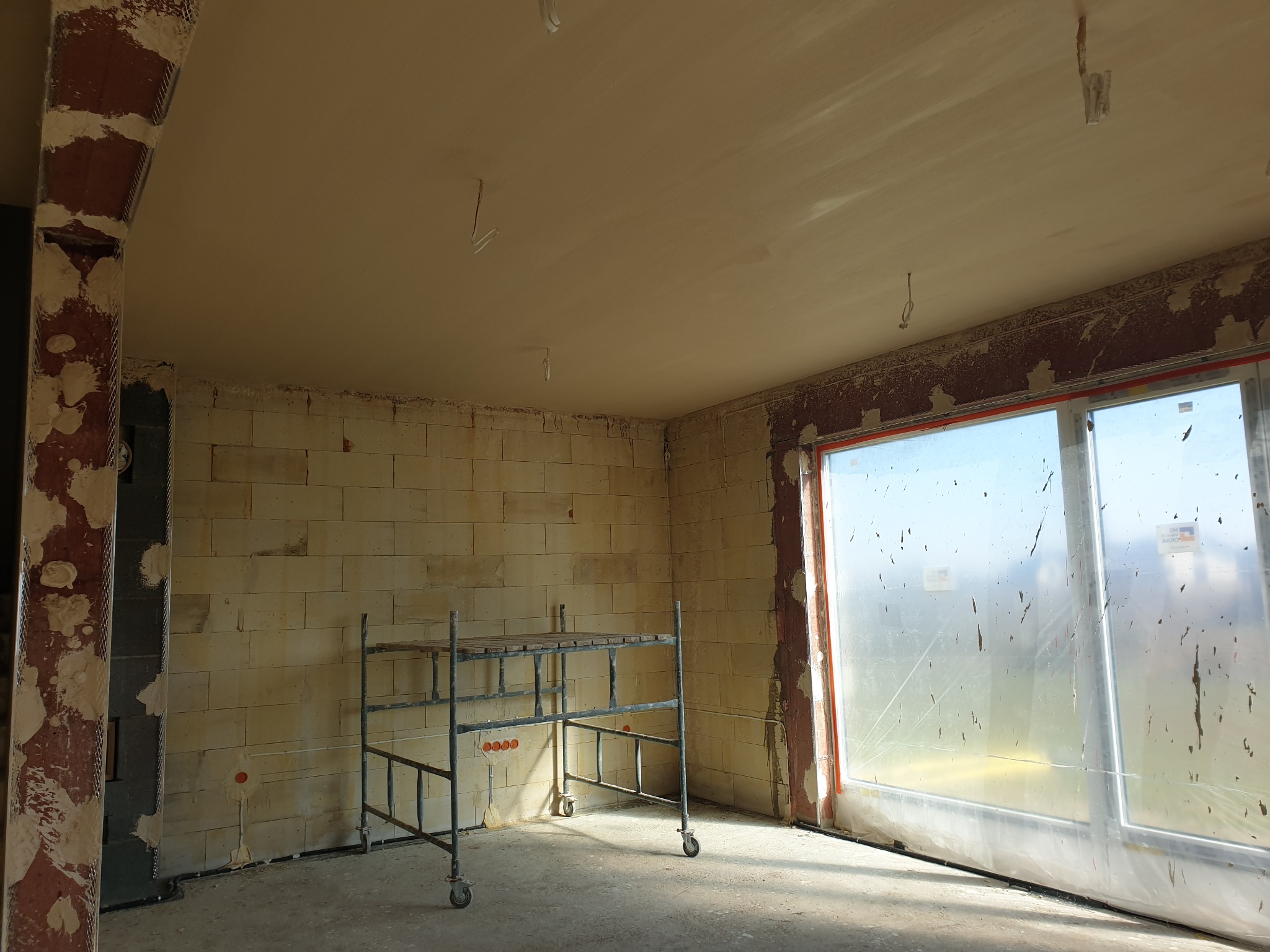 Wnętrze z nałożonymi tynkami, widoczne rusztowanie i okno zabezpieczone folią. Ściany z bloczków i wykończony sufit. Etap prac wykończeniowych.