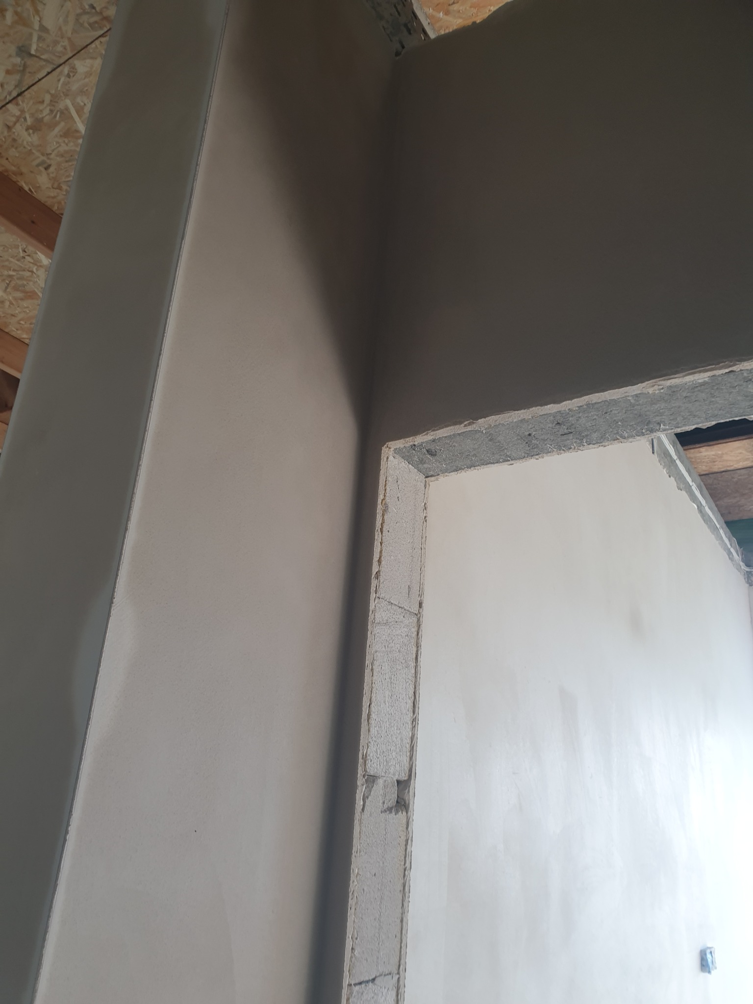 Świeżo nałożony, gładki jasnoszary tynk na ścianie i futrynie drzwiowej, widoczne ślady po pacie, sufit z płyt OSB w tle. Ukazany fragment wnętrza w trakcie prac wykończeniowych.