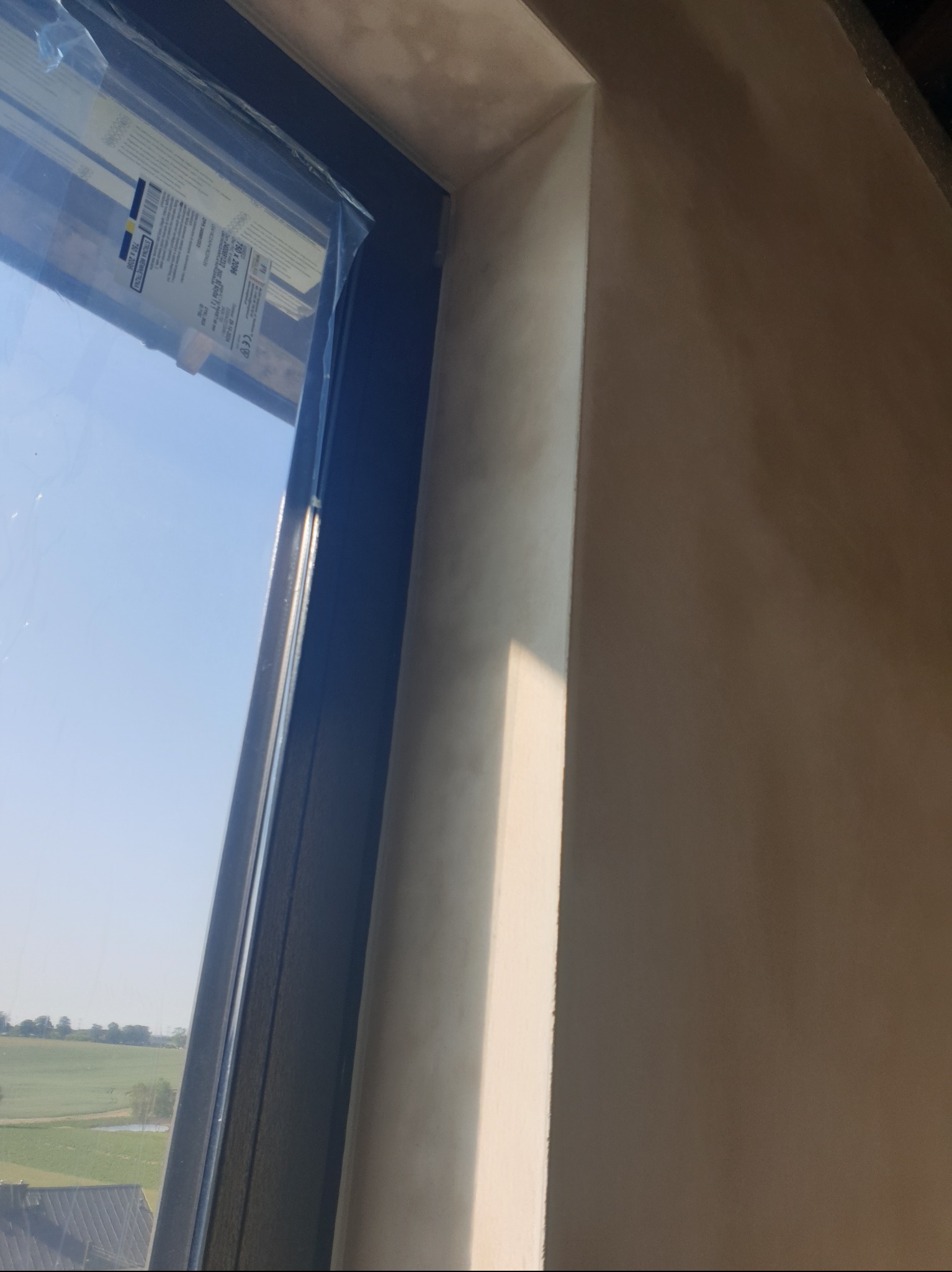 Gładka, beżowa ściana po tynkowaniu, widoczna obok okna z widokiem na zielone pola. Świeżo położony tynk z delikatną fakturą w naturalnym świetle.