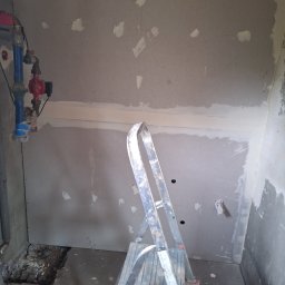 Bartosz Czerwiński - Ściana z płyt gipsowo-kartonowych z widocznymi miejscami po szpachlowaniu, fragment instalacji wodnej i drabina aluminiowa w pomieszczeniu w trakcie prac remontowych.