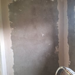 Bartosz Czerwiński - Ściana w trakcie prac przygotowawczych pod malowanie, widoczna nałożona warstwa szpachli w odcieniach szarości i beżu, nierównomiernie rozprowadzona, z widocznymi śladami narzędzi, w tle fragment...