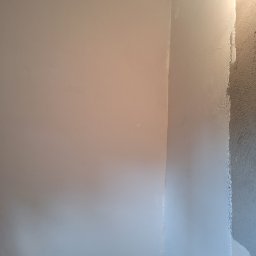 Bartosz Czerwiński - Róg pokoju w trakcie prac wykończeniowych: fragment ściany z nałożoną gładzią, obok fragment ściany z surowym tynkiem, widoczne otwory na gniazdka elektryczne.