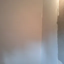 Róg pokoju w trakcie prac wykończeniowych: fragment ściany z nałożoną gładzią, obok fragment ściany z surowym tynkiem, widoczne otwory na gniazdka elektryczne.