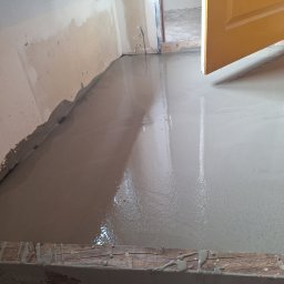 Bartosz Czerwiński - Świeżo wylana, mokra, samopoziomująca wylewka podłogowa w pomieszczeniu z otwartymi, żółtymi drzwiami, widoczne ślady zaprawy na ścianie i drewniana listwa przypodłogowa z resztkami wylewki.