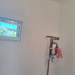 Bartosz Czerwiński - Wnętrze w trakcie prac wykończeniowych: widoczne białe ściany, okno z widokiem na zieleń oraz instalacja centralnego ogrzewania z zawieszonym ręcznikiem.