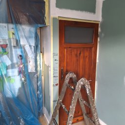 Bartosz Czerwiński - Przygotowania do malowania ściany: drewniane drzwi zabezpieczone taśmą malarską, drabina oparta o ścianę, meble osłonięte folią, fragment ściany pomalowany na zielono, widoczne ślady po malowaniu...