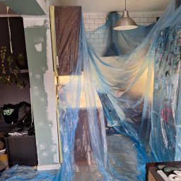 Bartosz Czerwiński - Przygotowanie do malowania mieszkania: zabezpieczone folią meble i podłoga, ściany z widocznymi śladami po szpachlowaniu, sufit z lampą, widoczne narzędzia malarskie.