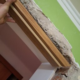 Bartosz Czerwiński - Mężczyzna przykłada drewnianą ościeżnicę do surowego otworu drzwiowego z widocznymi cegłami i resztkami tynku na zielonej ścianie.