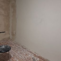 Bartosz Czerwiński - Pokój w trakcie szpachlowania ścian, widoczna częściowo wyszpachlowana ściana, resztki szpachli na podłodze i szpachelka leżąca na podłodze, mały stołek obok ściany.