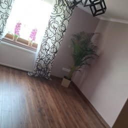 Bartosz Czerwiński - Pokój z drewnianą podłogą, okno z wzorzystymi zasłonami, kwiaty na parapecie, roślina doniczkowa w rogu i nowoczesna lampa sufitowa z trzema kwadratowymi kloszami.