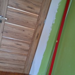 Bartosz Czerwiński - Wnętrze w trakcie prac: drewnopodobne panele podłogowe, drewniana drzwi z poziomymi srebrnymi wstawkami, zielona ściana częściowo pokryta białą masą szpachlową i czerwona poziomica oparta o ścianę.