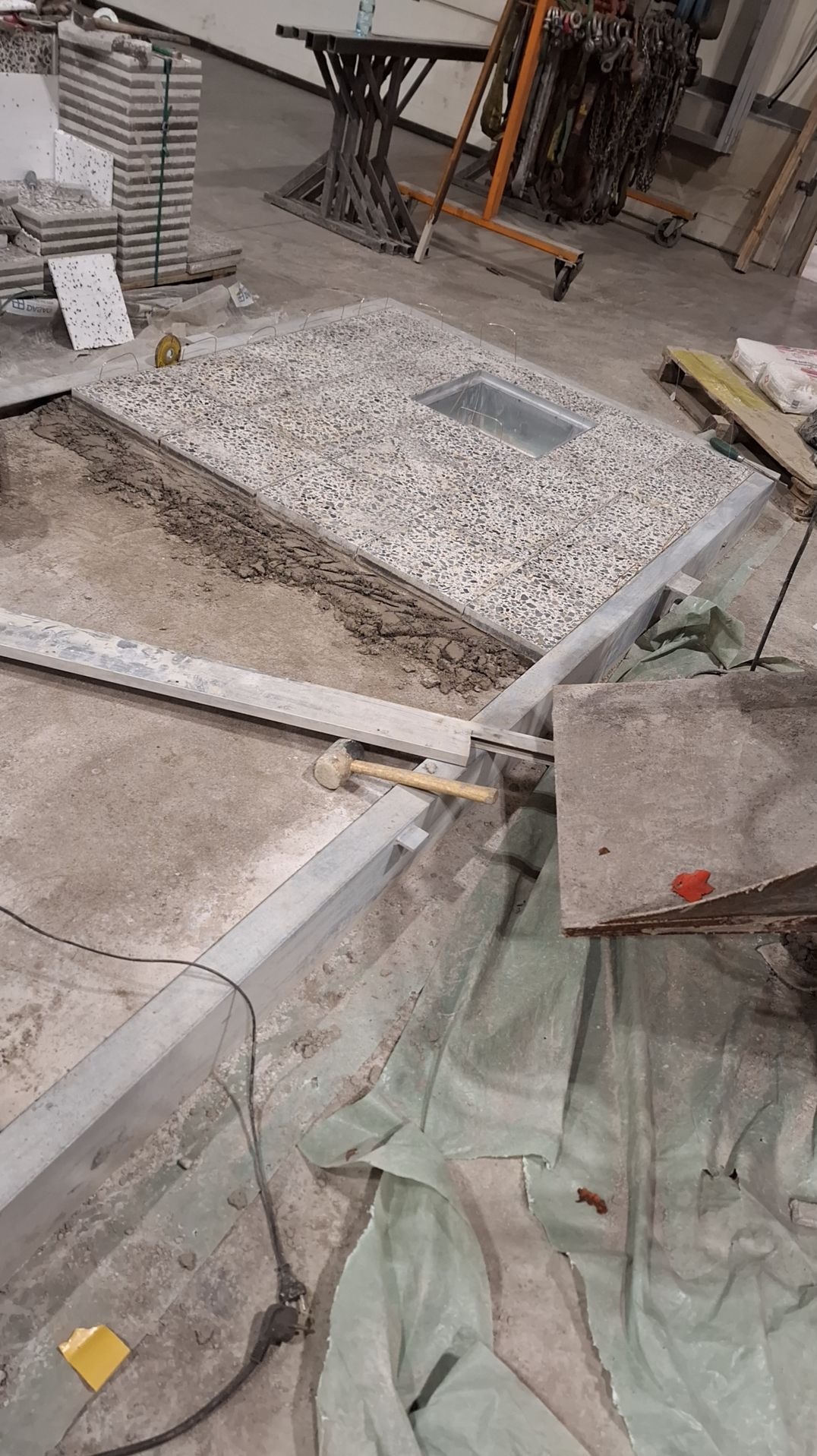 Fragment podłogi z mozaikowych płytek, z widocznym otworem wentylacyjnym, otoczony metalową ramą, gumowy młotek leżący na ramie, obok stos płytek i narzędzia.