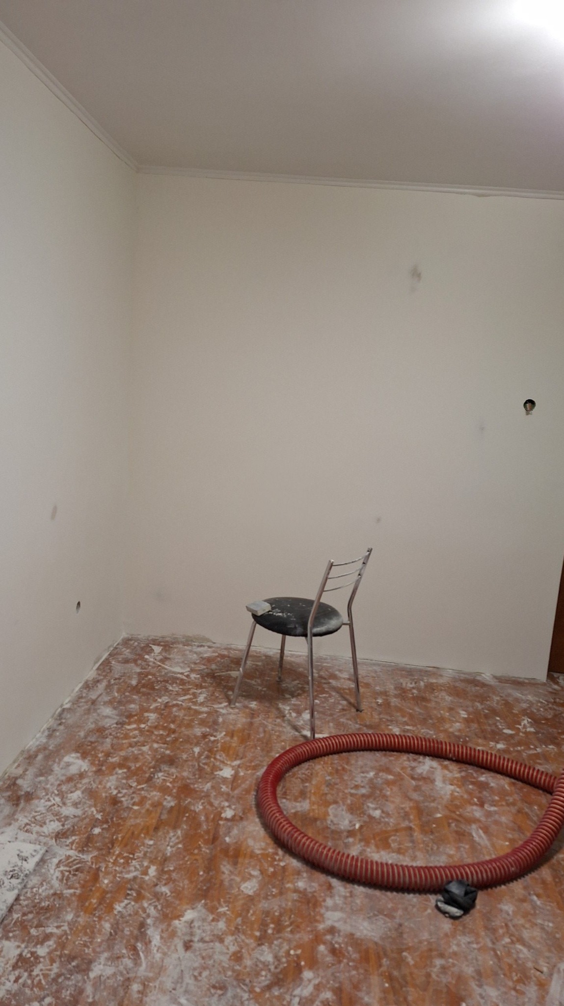 Pokój w trakcie przygotowania do prac wykończeniowych: ściany po szpachlowaniu, podłoga zabezpieczona folią i pokryta warstwą pyłu, czerwony wąż od odkurzacza przemysłowego i krzesło pośrodku...