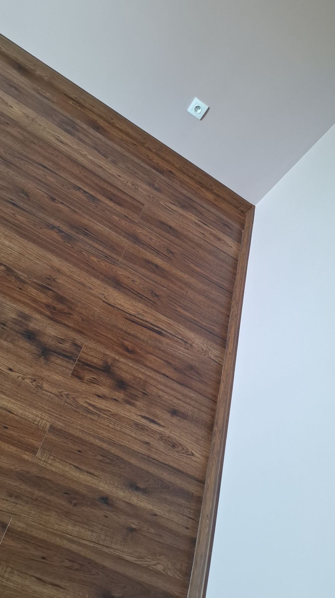 Fragment ściany wykończonej panelami drewnopodobnymi w odcieniach brązu, stykającej się z białą ścianą i sufitem, widoczne gniazdko elektryczne.