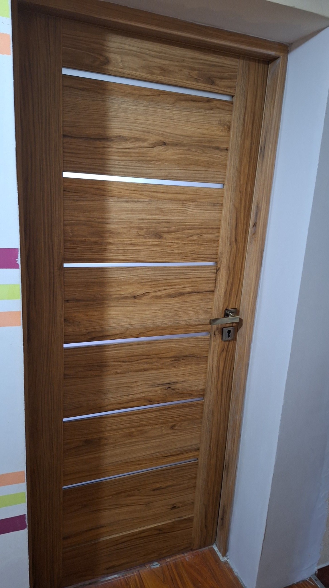 Drewniane drzwi wewnętrzne z poziomymi, srebrnymi wstawkami, zamontowane w białej ścianie, widoczna klamka i fragment podłogi.