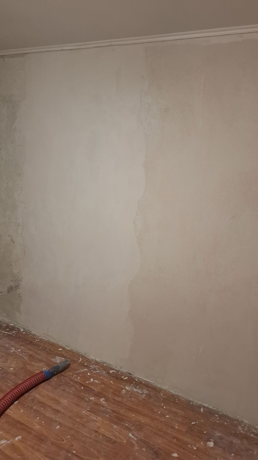 Ściana w trakcie prac przygotowawczych, częściowo pokryta gładzią, widoczny wąż od odkurzacza i zabrudzona podłoga z paneli.