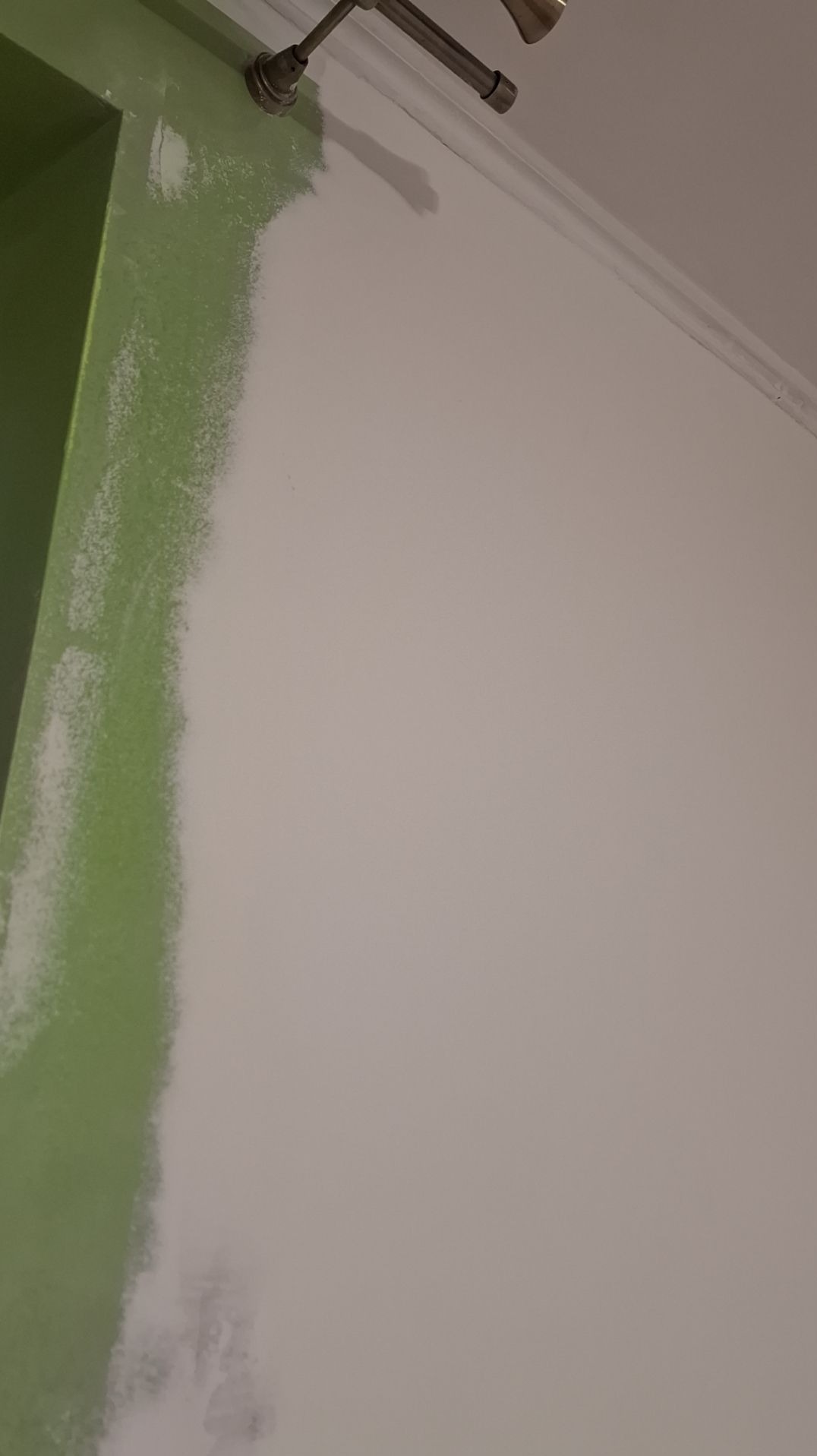 Ściana z fragmentem pomalowanej na zielono framugi drzwi, częściowo pokryta świeżą warstwą białej farby, widoczna listwa sufitowa i fragment karnisza.
