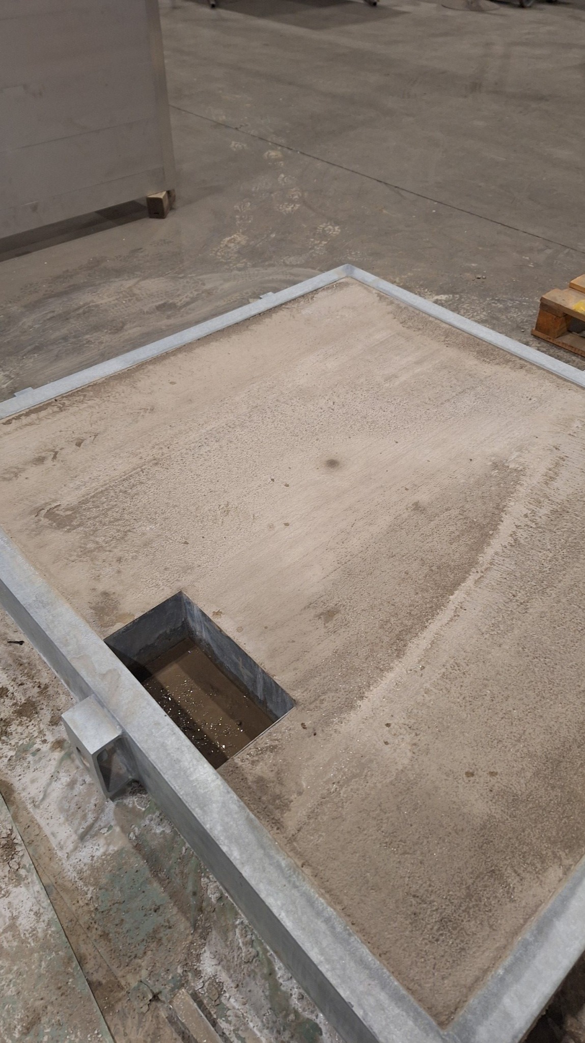 Szara, kwadratowa konstrukcja z otworem wypełnionym wodą, widoczna na szorstkiej, betonowej podłodze w hali.