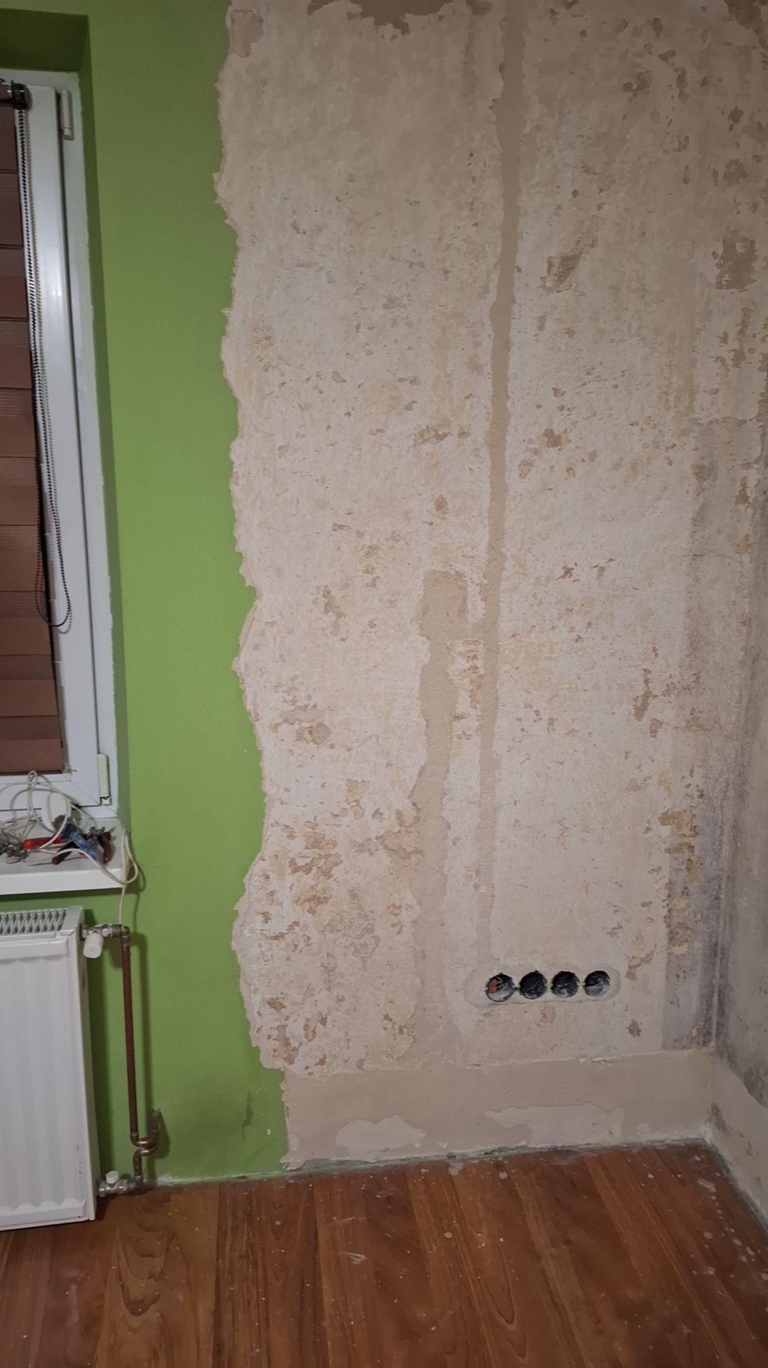 Ściana w trakcie przygotowania do prac wykończeniowych: fragment pomalowany na zielono, reszta z resztkami starej farby i widocznymi gniazdkami elektrycznymi, drewnopodobna podłoga.
