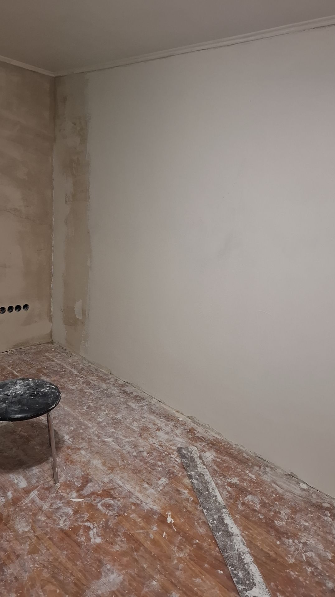 Pokój w trakcie szpachlowania ścian, widoczna częściowo wyszpachlowana ściana, resztki szpachli na podłodze i szpachelka leżąca na podłodze, mały stołek obok ściany.