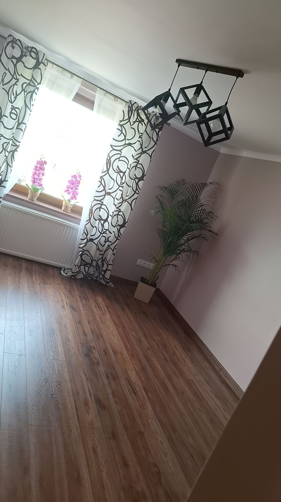 Pokój z drewnianą podłogą, okno z wzorzystymi zasłonami, kwiaty na parapecie, roślina doniczkowa w rogu i nowoczesna lampa sufitowa z trzema kwadratowymi kloszami.