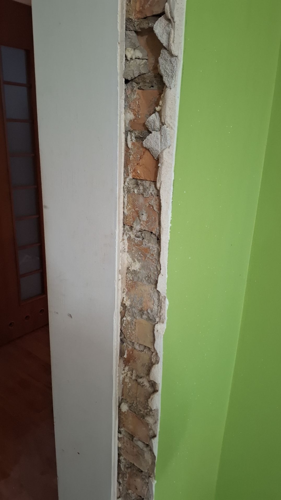 Odsłonięta cegła i resztki zaprawy w ościeżnicy drzwiowej, widoczne przygotowania do osadzania futryny na tle zielonej ściany.