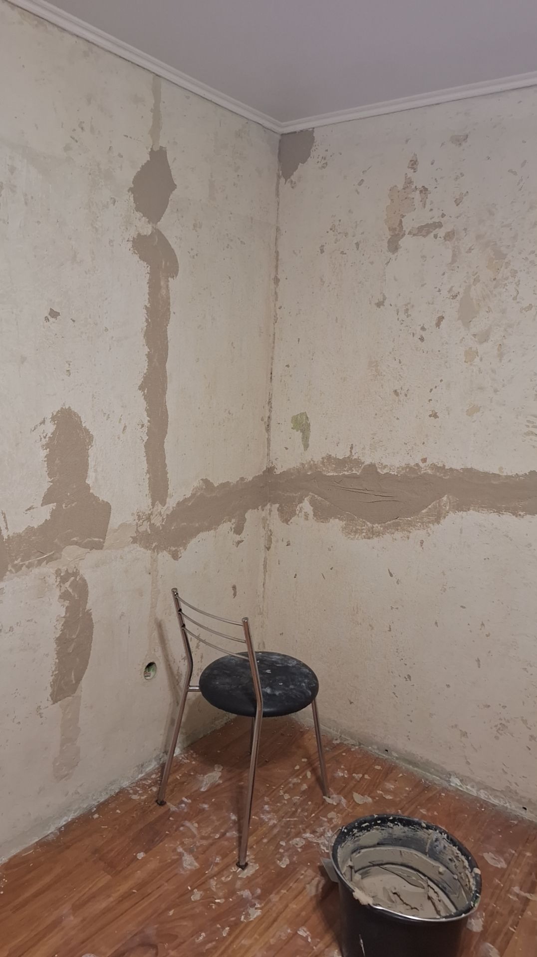 Przygotowanie ścian do dalszych prac wykończeniowych, widoczne nałożone warstwy szpachli w narożnikach i wzdłuż ściany, krzesło i wiadro z zaprawą na podłodze z panelami.