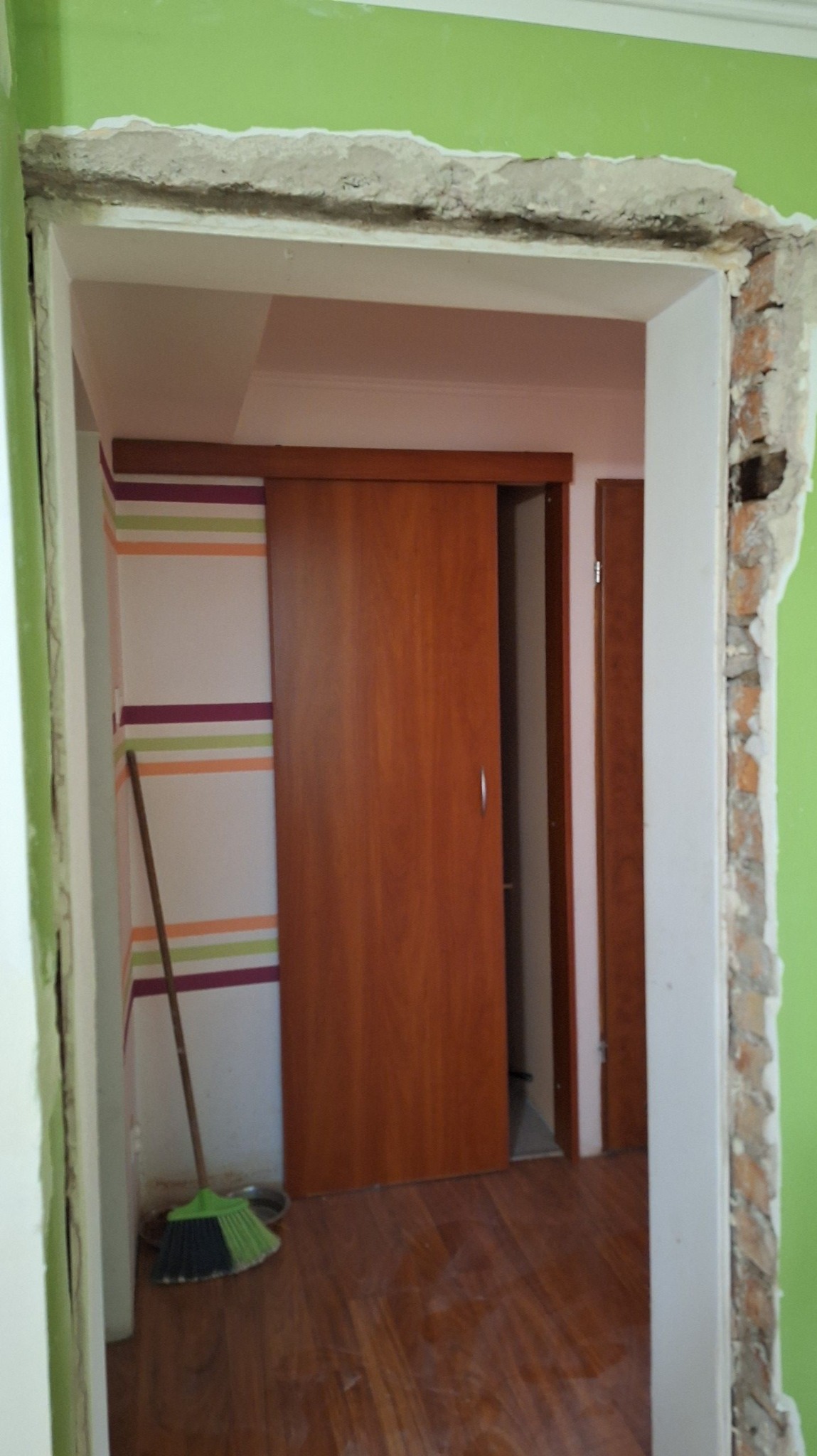 Montaż drzwi przesuwnych w trakcie remontu, widoczne uszkodzone ściany i listwy, podłoga drewniana, ściana w paski, zielona ściana, miotła w kącie.