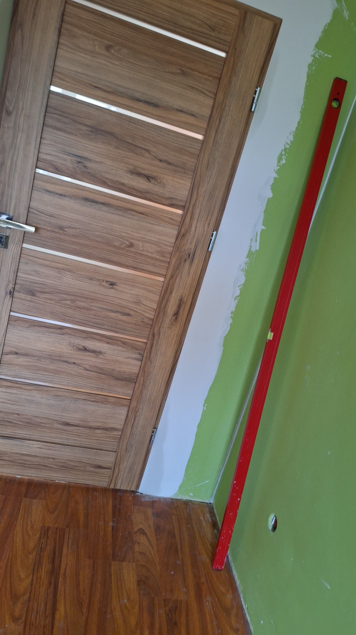 Wnętrze w trakcie prac: drewnopodobne panele podłogowe, drewniana drzwi z poziomymi srebrnymi wstawkami, zielona ściana częściowo pokryta białą masą szpachlową i czerwona poziomica oparta o ścianę.