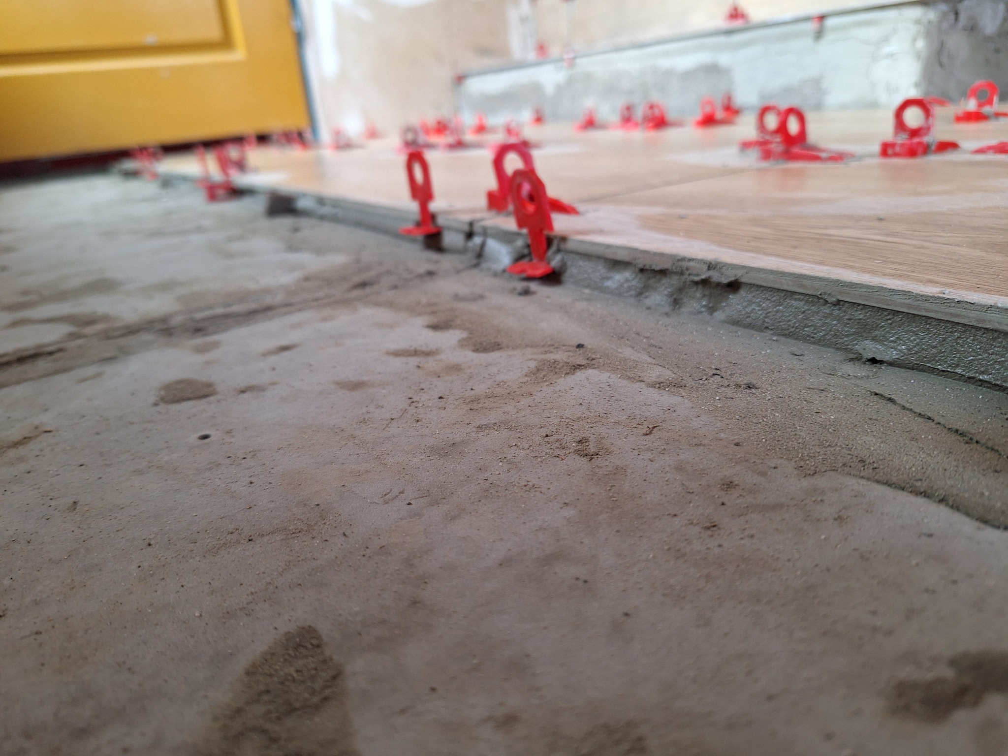 Układanie płytek podłogowych w Bledzewie z użyciem czerwonych klipsów poziomujących, widoczna warstwa zaprawy i fragment żółtych drzwi w tle.