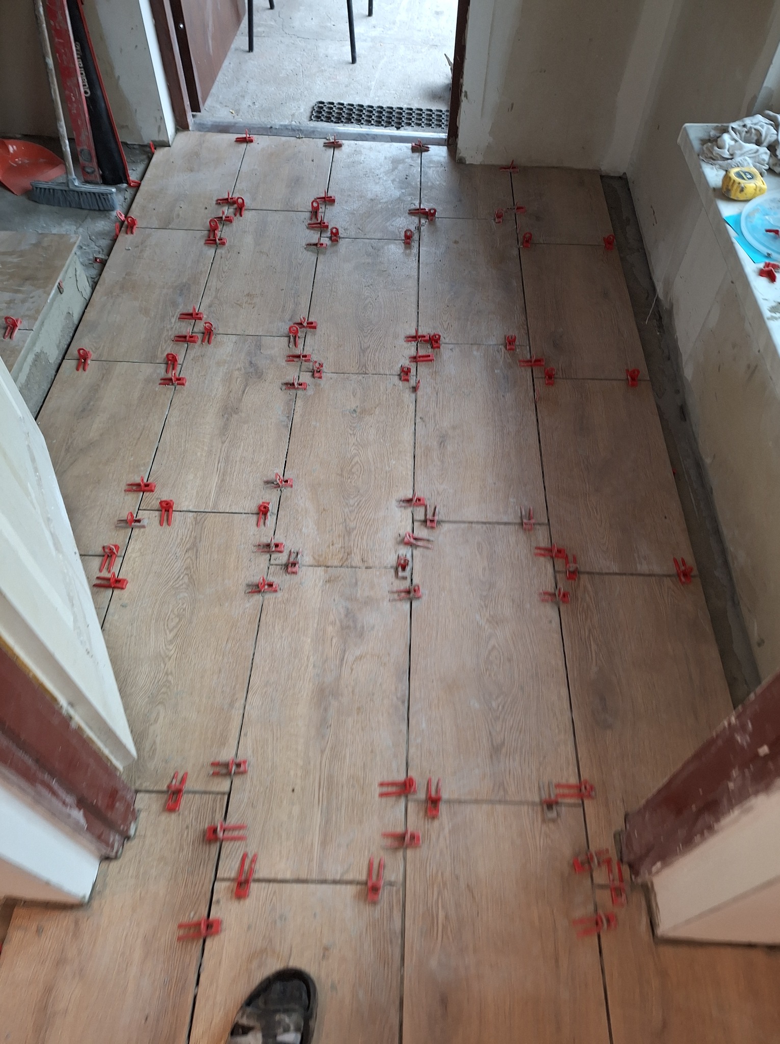 Podłoga z płytek imitujących drewno w trakcie układania, z użyciem czerwonych klipsów poziomujących, widoczny fragment buta w dolnym rogu.