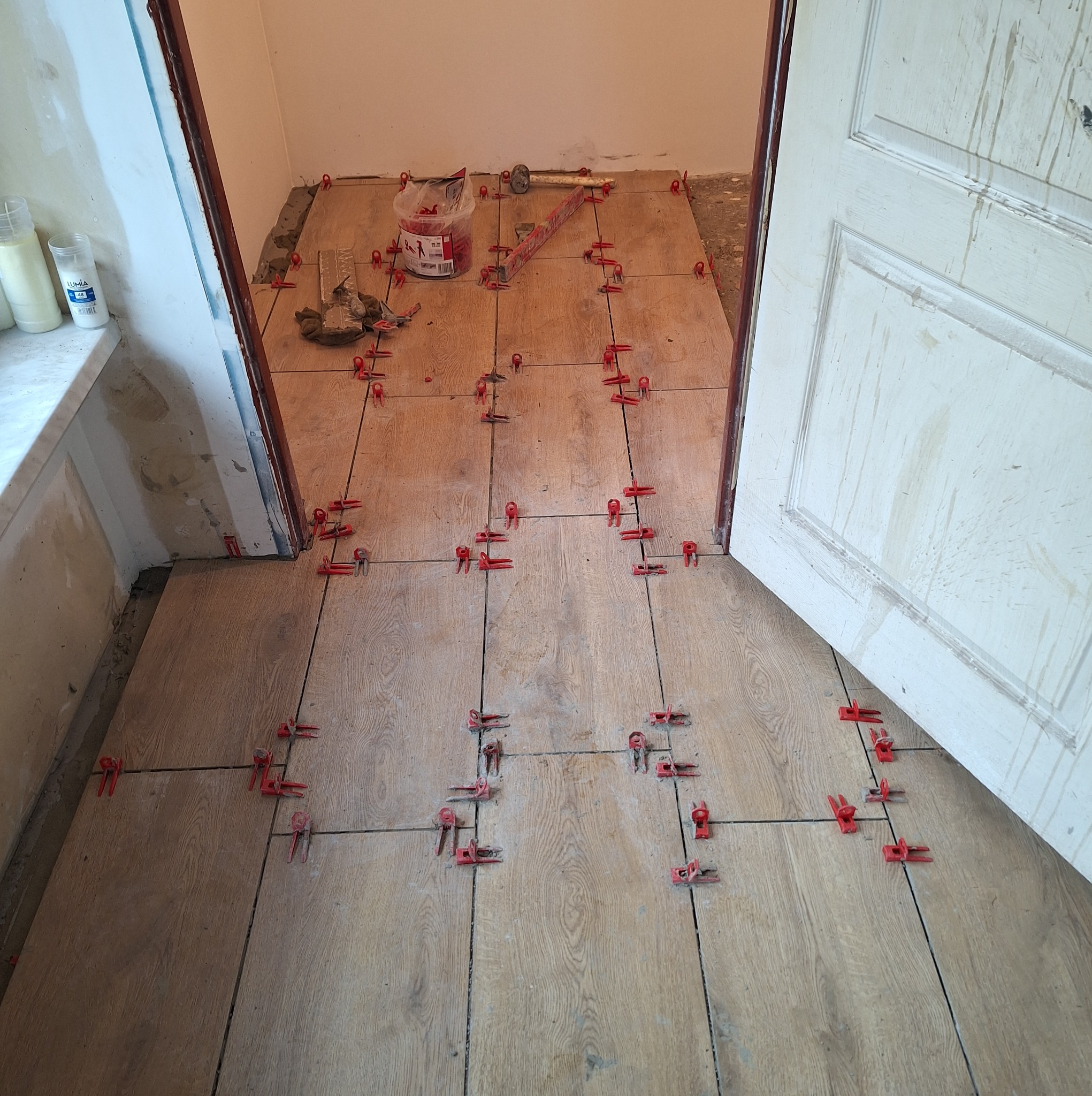 Ułożone płytki podłogowe imitujące drewno z czerwonymi klipsami poziomującymi, widoczny fragment drzwi i narzędzia glazurnicze w tle.