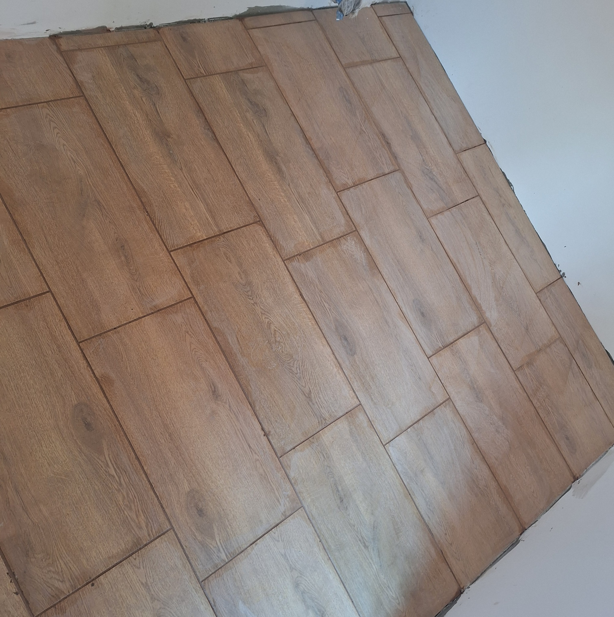 Ułożone płytki podłogowe imitujące drewno w rogu pomieszczenia, widoczne niedoskonałości przy ścianie przed fugowaniem.