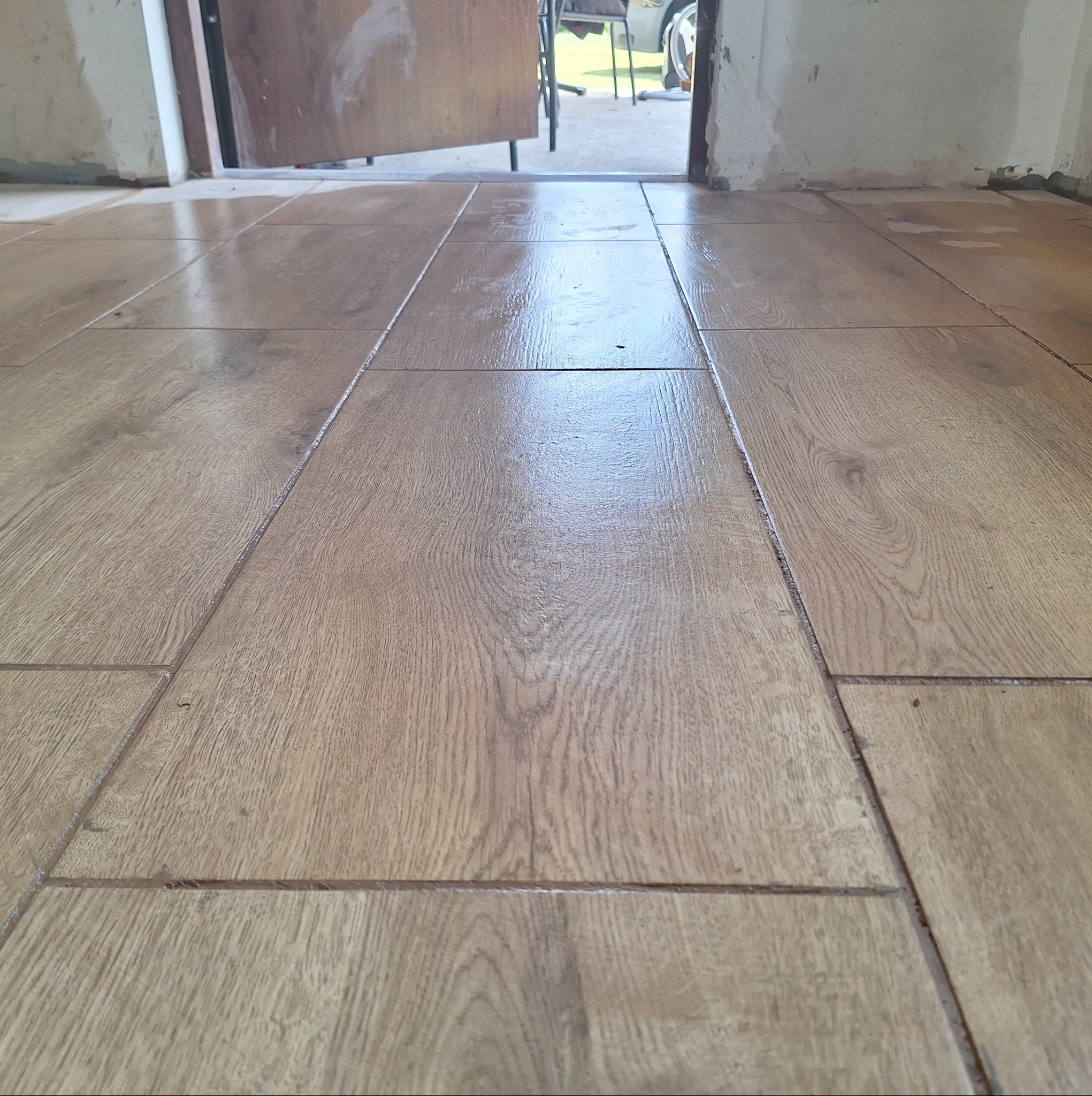 Świeżo ułożone, błyszczące panele podłogowe imitujące drewno, widoczne fugi, otwarte drzwi w tle.