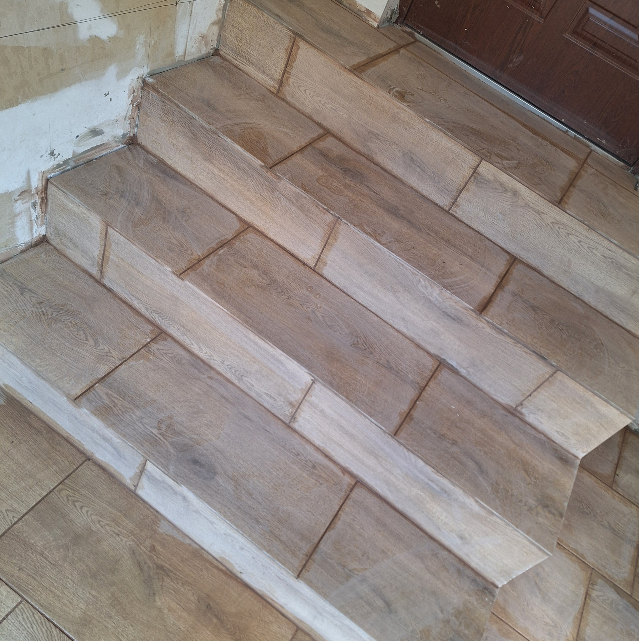 Wykończone schody z płytek imitujących drewno, z widocznymi śladami po pracach wykończeniowych na ścianie i przy futrynie.