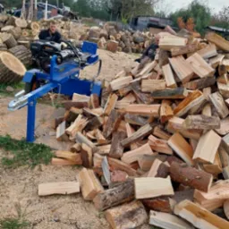 Stos drewna kominkowego po obróbce, z łuparką hydrauliczną w tle, otoczone wiórami. Widoczny mężczyzna obsługujący maszynę. Sezon jesienny.