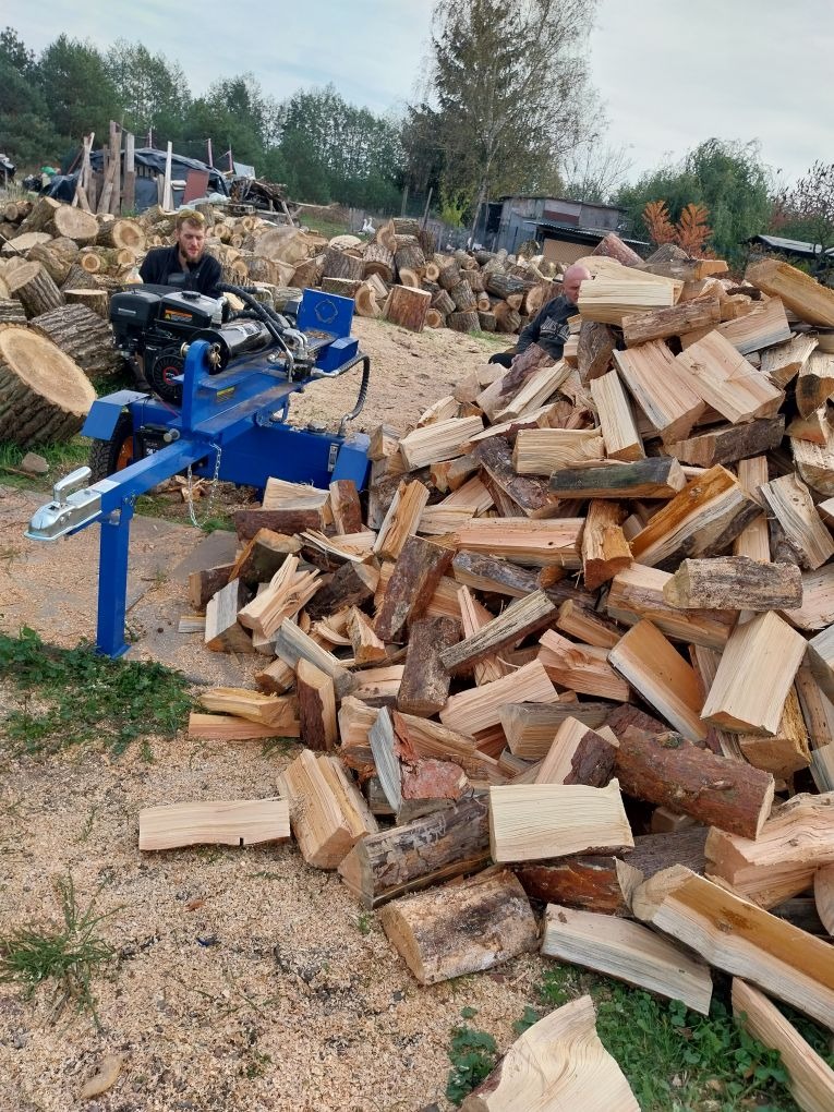 Stos drewna kominkowego po obróbce, z łuparką hydrauliczną w tle, otoczone wiórami. Widoczny mężczyzna obsługujący maszynę. Sezon jesienny.