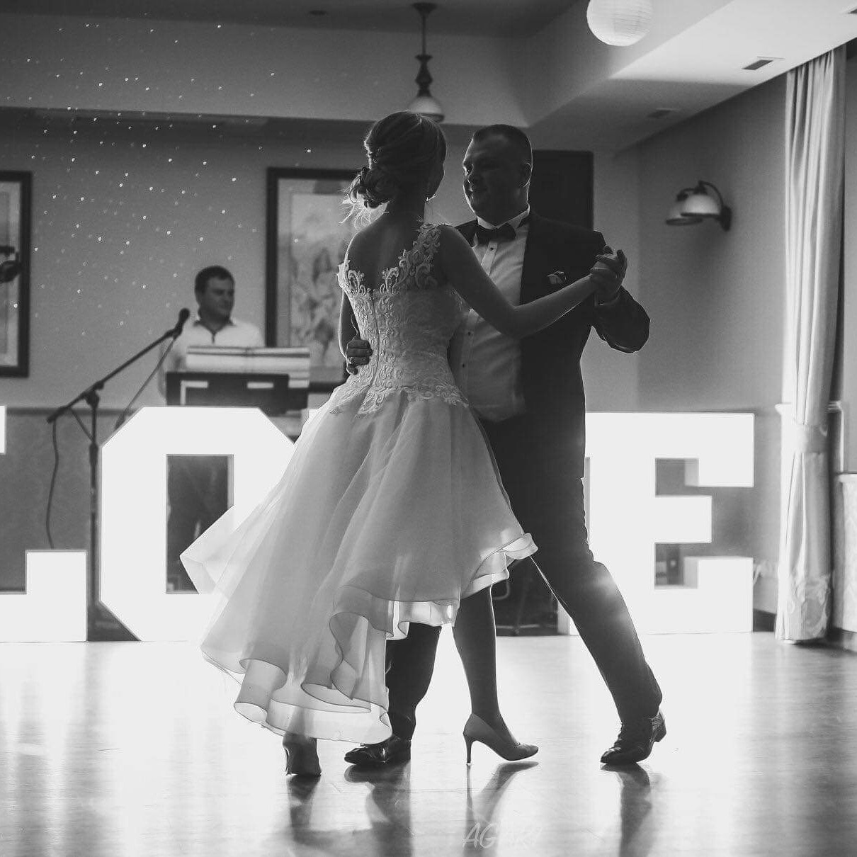 Czarno-białe zdjęcie tańczącej pary młodej w sukni ślubnej z asymetrycznym dołem i koronkowym gorsetem, obok podświetlonego napisu LOVE, w tle muzyk przy keyboardzie.