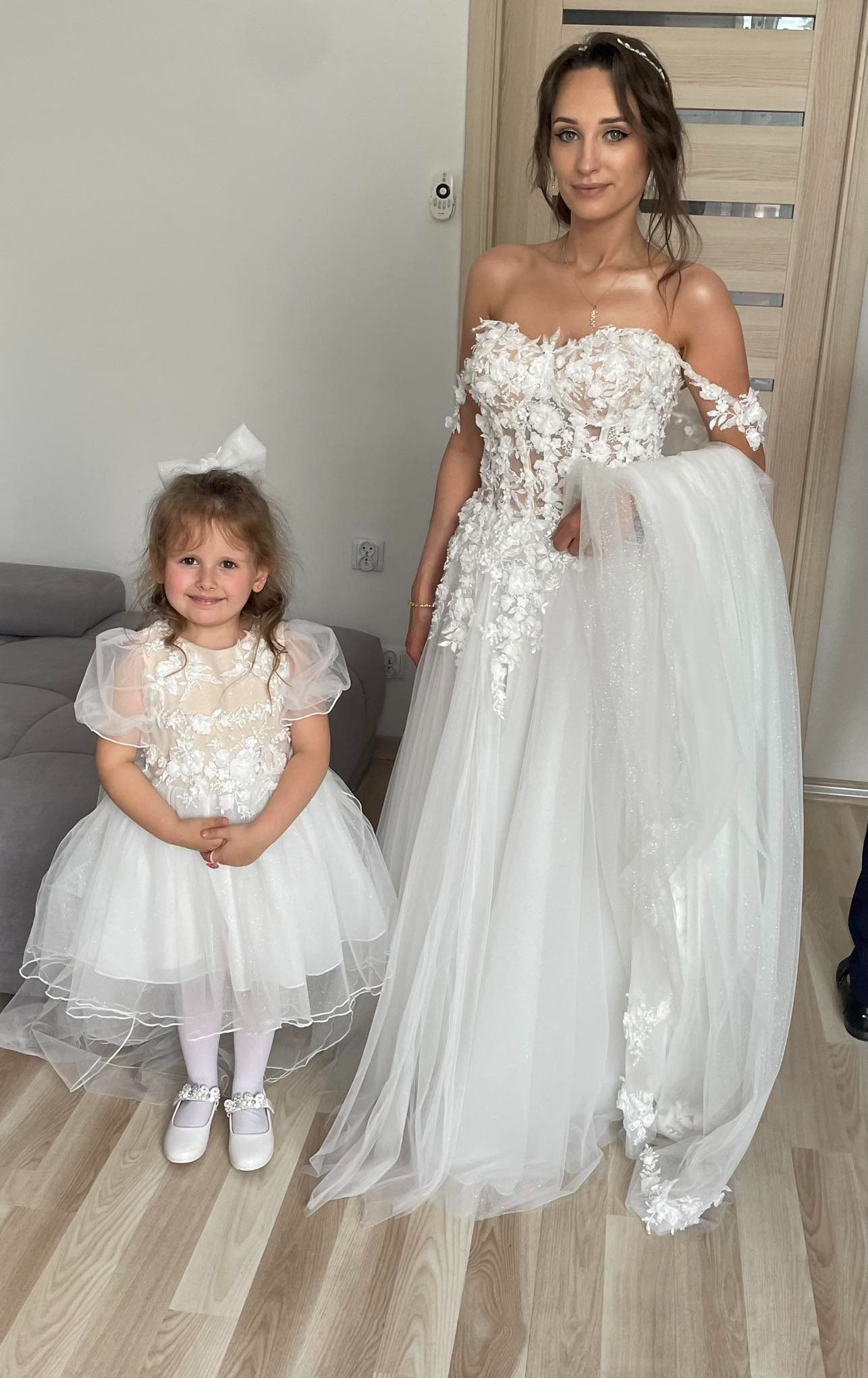 Panna młoda w białej sukni ślubnej z koronkową górą i tiulowym dołem, pozująca obok uśmiechniętej dziewczynki w dopasowanej sukience z kokardą we włosach.